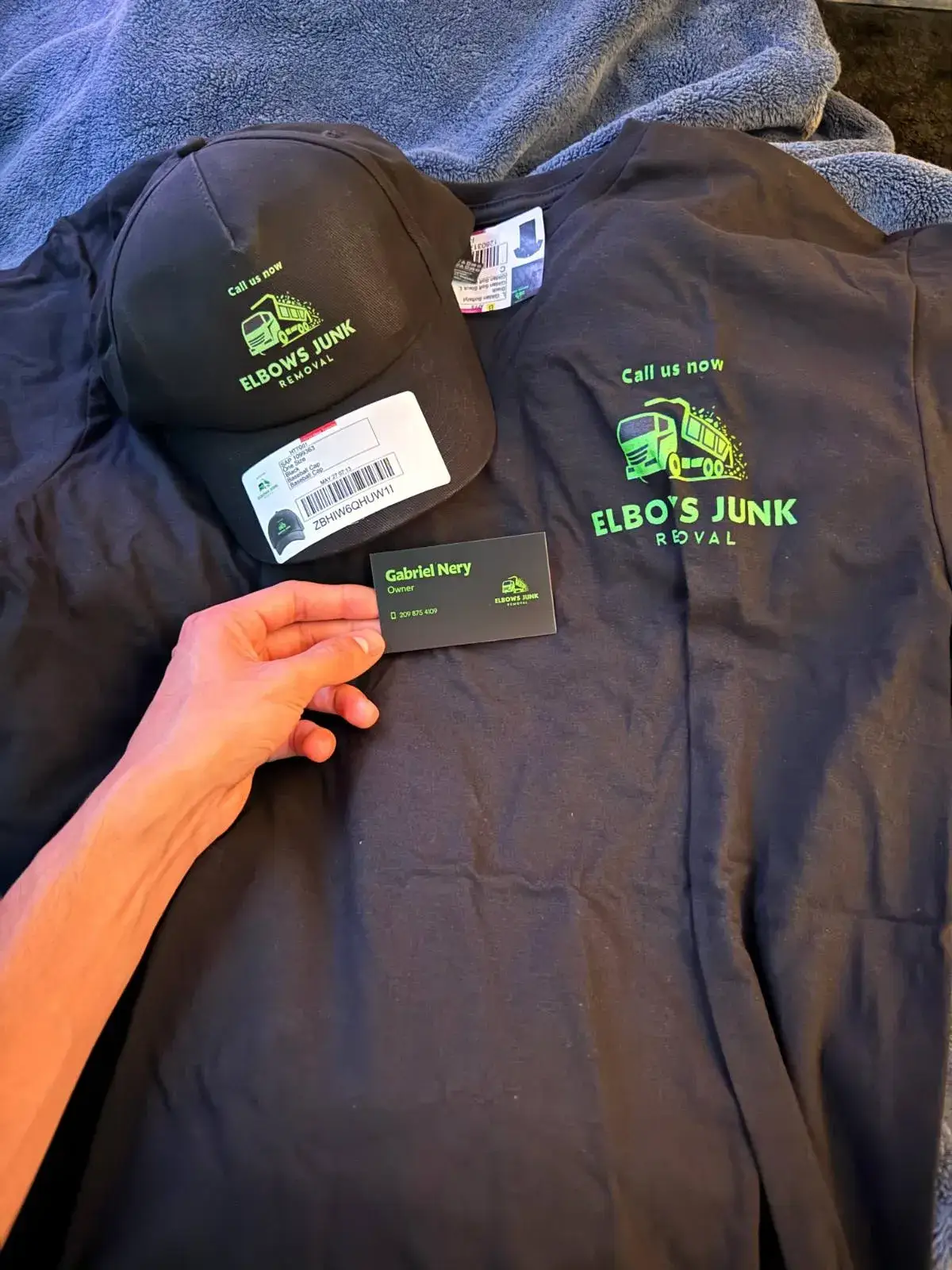 A hand holds an owner's business card above a black cap and shirt with bright green Elbows Junk Removal logos.