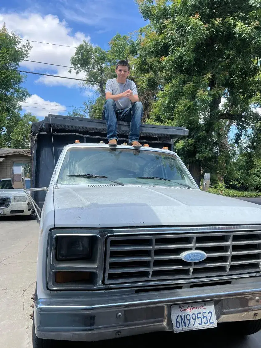 A person in a gray T-shirt and jeans sits above the cab of a silver Ford truck with a tall black cargo body, parked outdoors beneath trees and a blue sky.