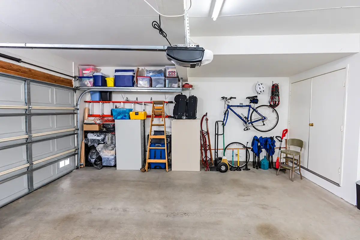 Garage interior with a clear concrete floor, storage shelves and cabinets, a wall-mounted bicycle, ladders, tools, and an overhead garage door opener.